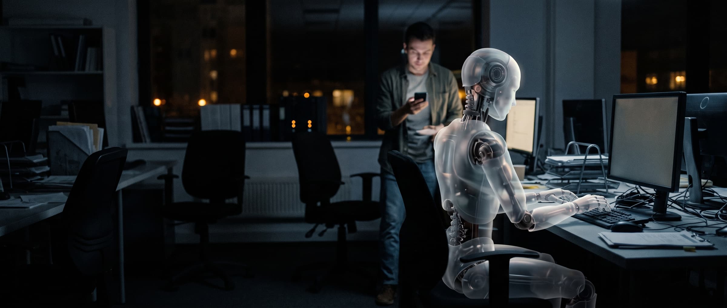 A translucent robot works at a terminal at night while a person behind it checks a phone to approve.