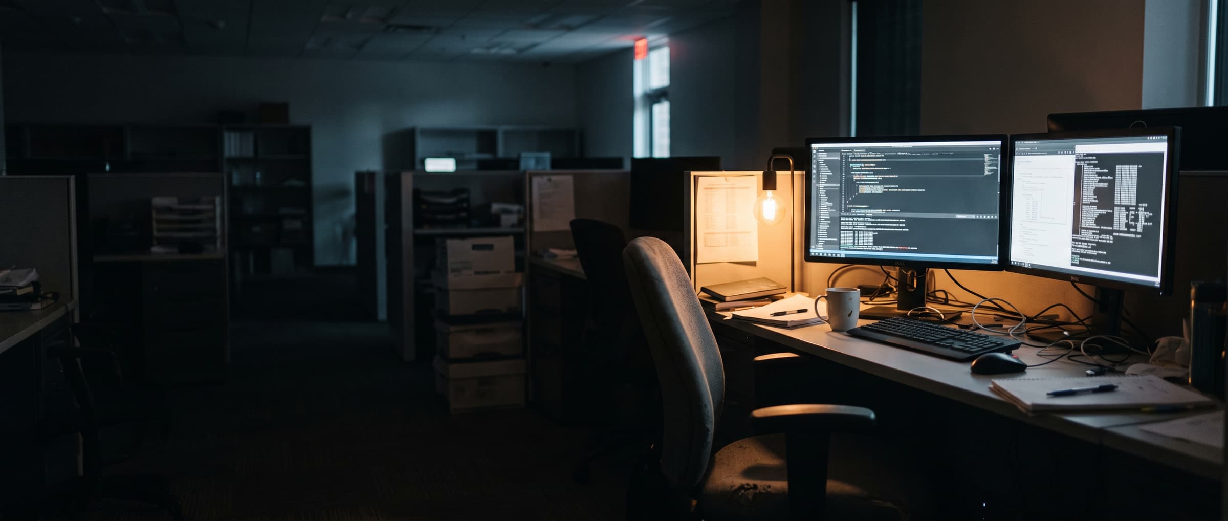 An empty desk at night, two monitors still running code with nobody in the chair.